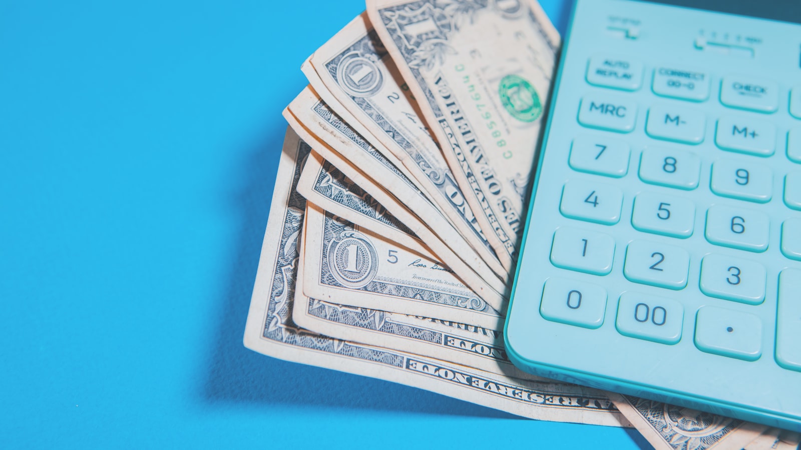 A teal calculator rests beside a fan of US dollar bills on a bright blue surface