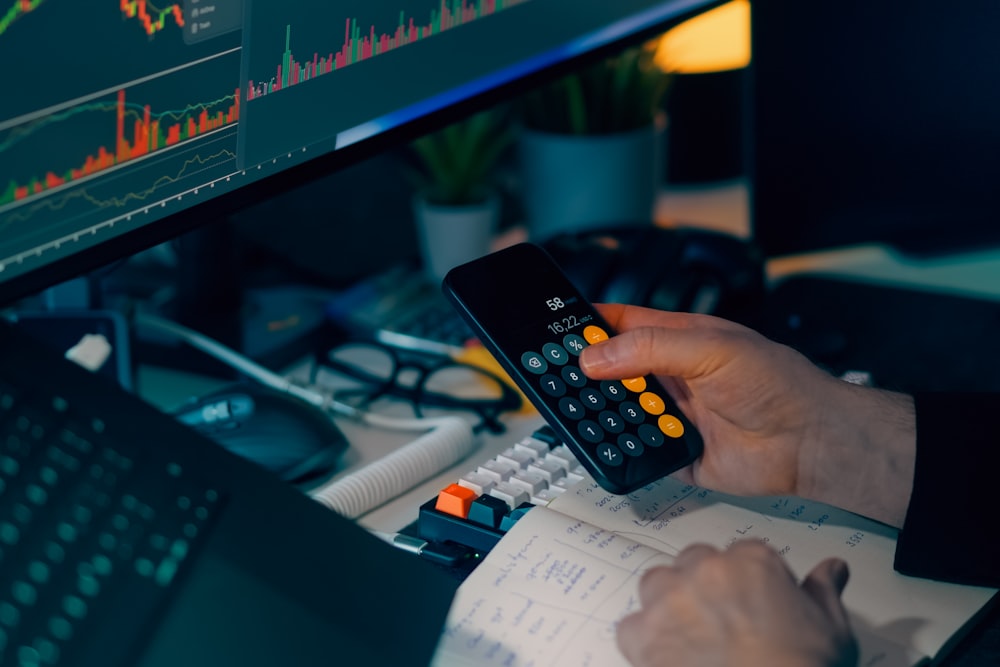 A person holds a phone displaying a calculator app over a desk with a laptop and a monitor showing a chart