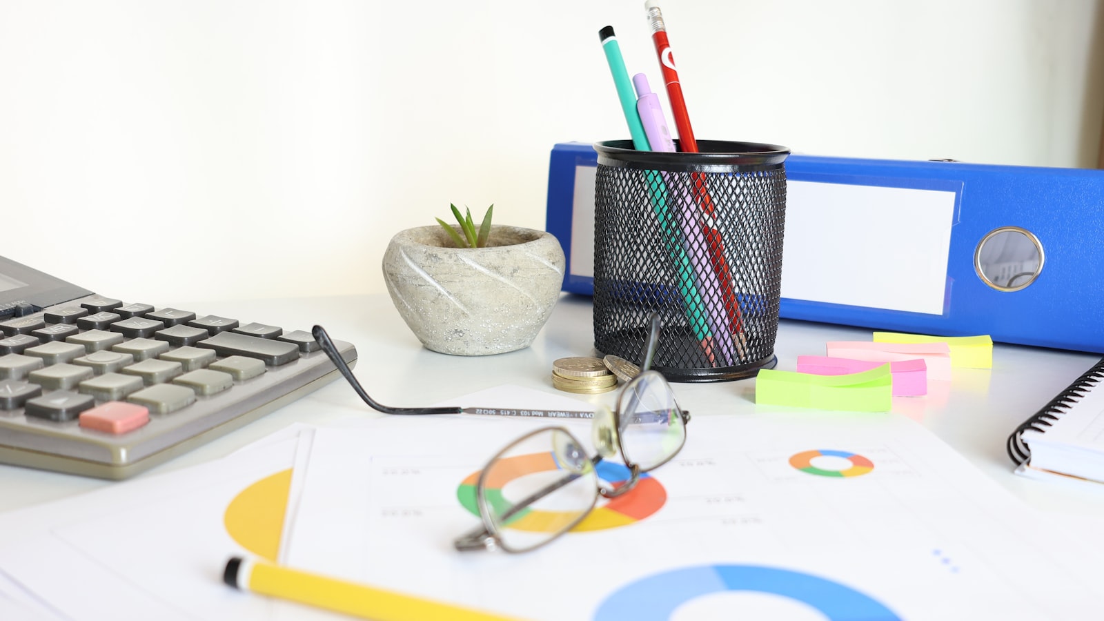 An office desk with a calculator, glasses, chart papers, pen holder, plant, and blue binder