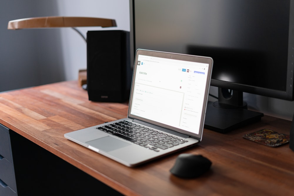An open laptop, a computer mouse, a monitor, a speaker, and a lamp on a wooden desk