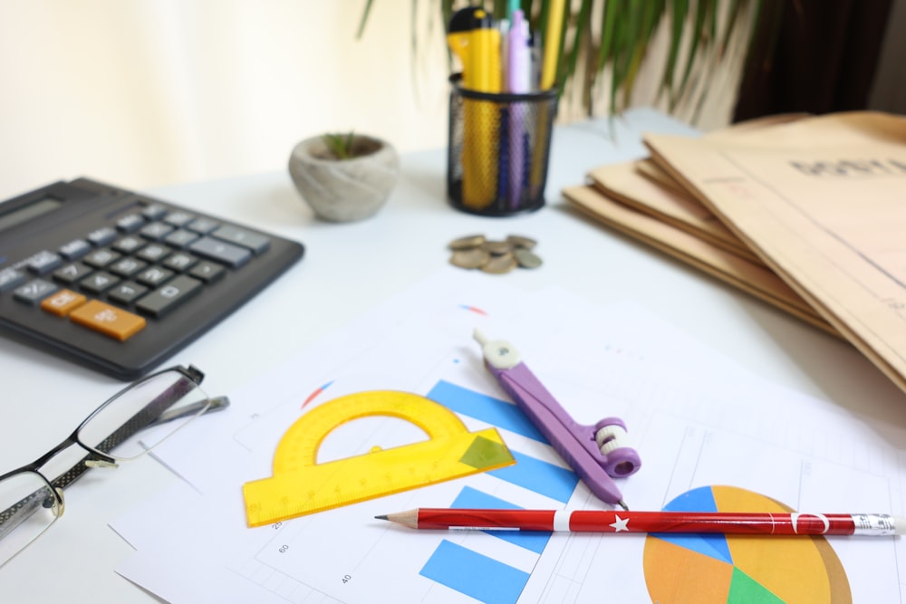 A desk with a calculator, eyeglasses, a protractor, compass, pencil, folders, and coins