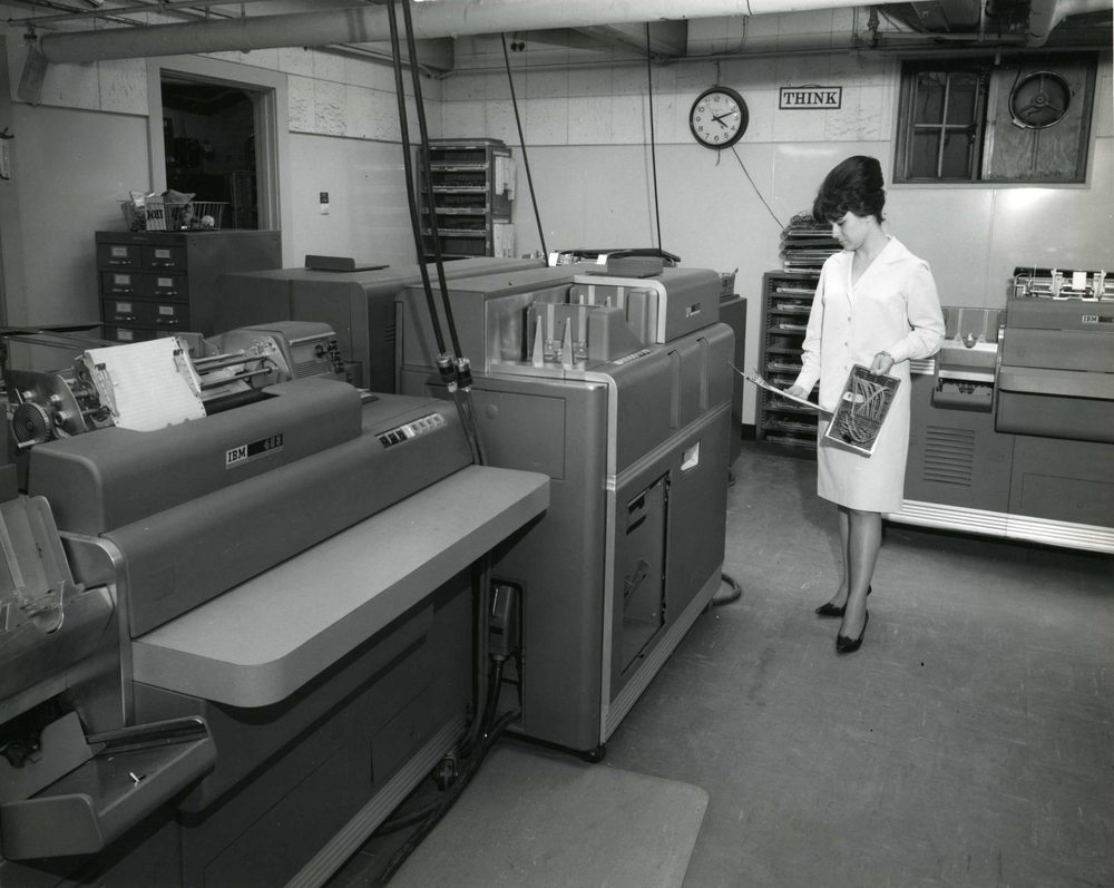 A woman in a lab coat examines a wired panel among large computers in an office