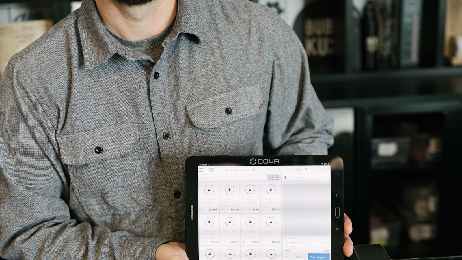 A man in a grey button-down shirt holds a tablet displaying a point-of-sale software interface