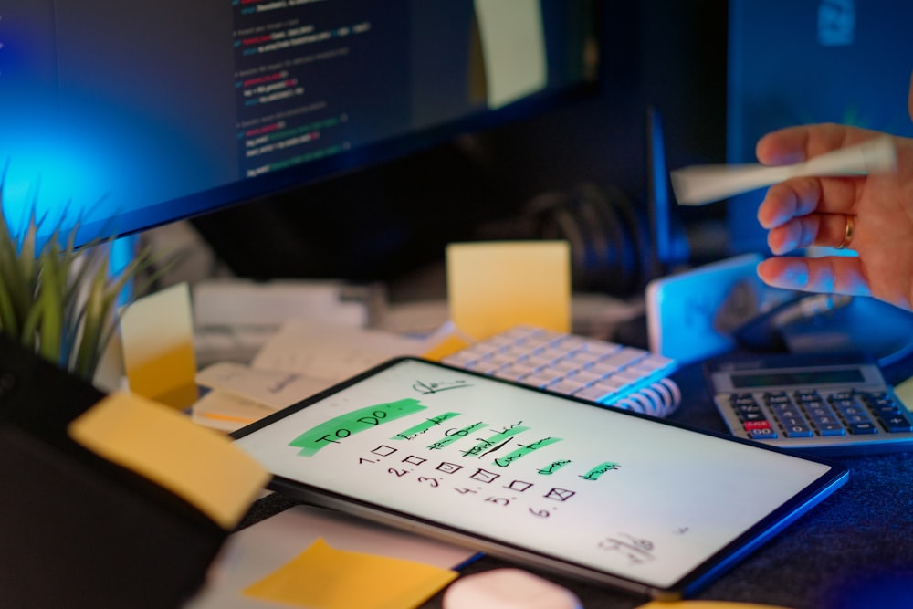 A tablet displaying a handwritten to-do list rests on a messy desk with a computer monitor, calculator, and sticky notes