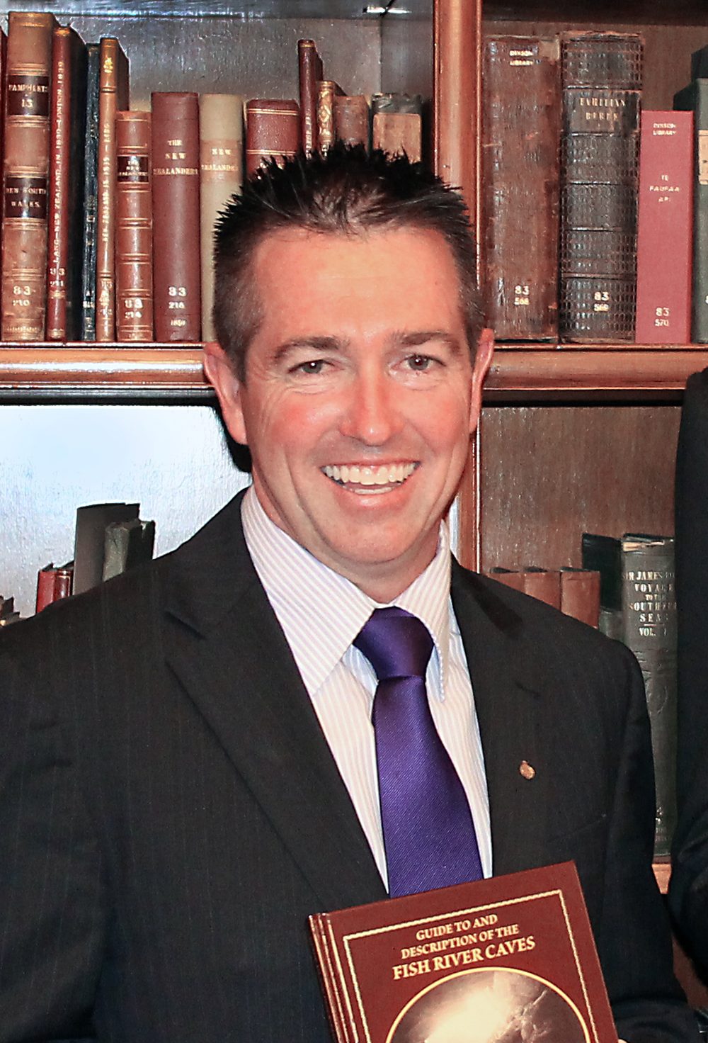 A smiling man in a suit holding a book in front of a bookshelf