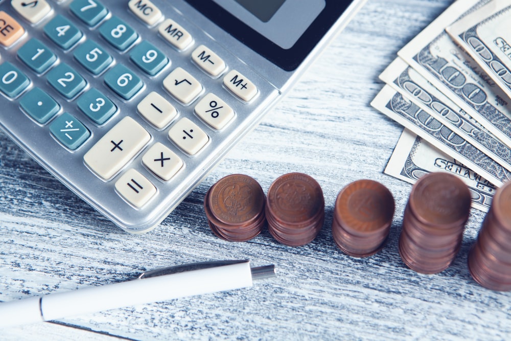 A calculator, stacks of coins, dollar bills, and a pen on a wooden table