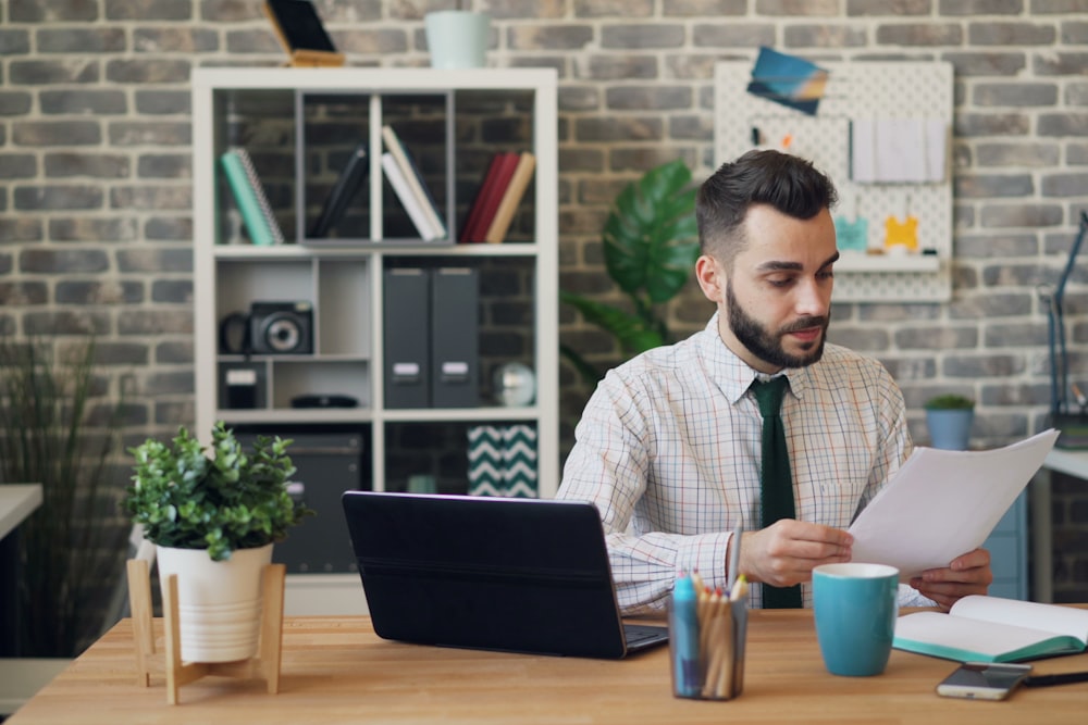 A bearded man sits at a wooden desk, holding papers and looking at them