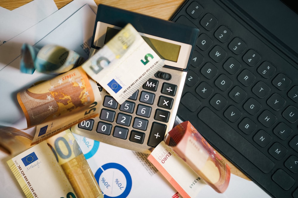 Euro banknotes, a calculator, papers, and a keyboard are arranged on a wooden desk