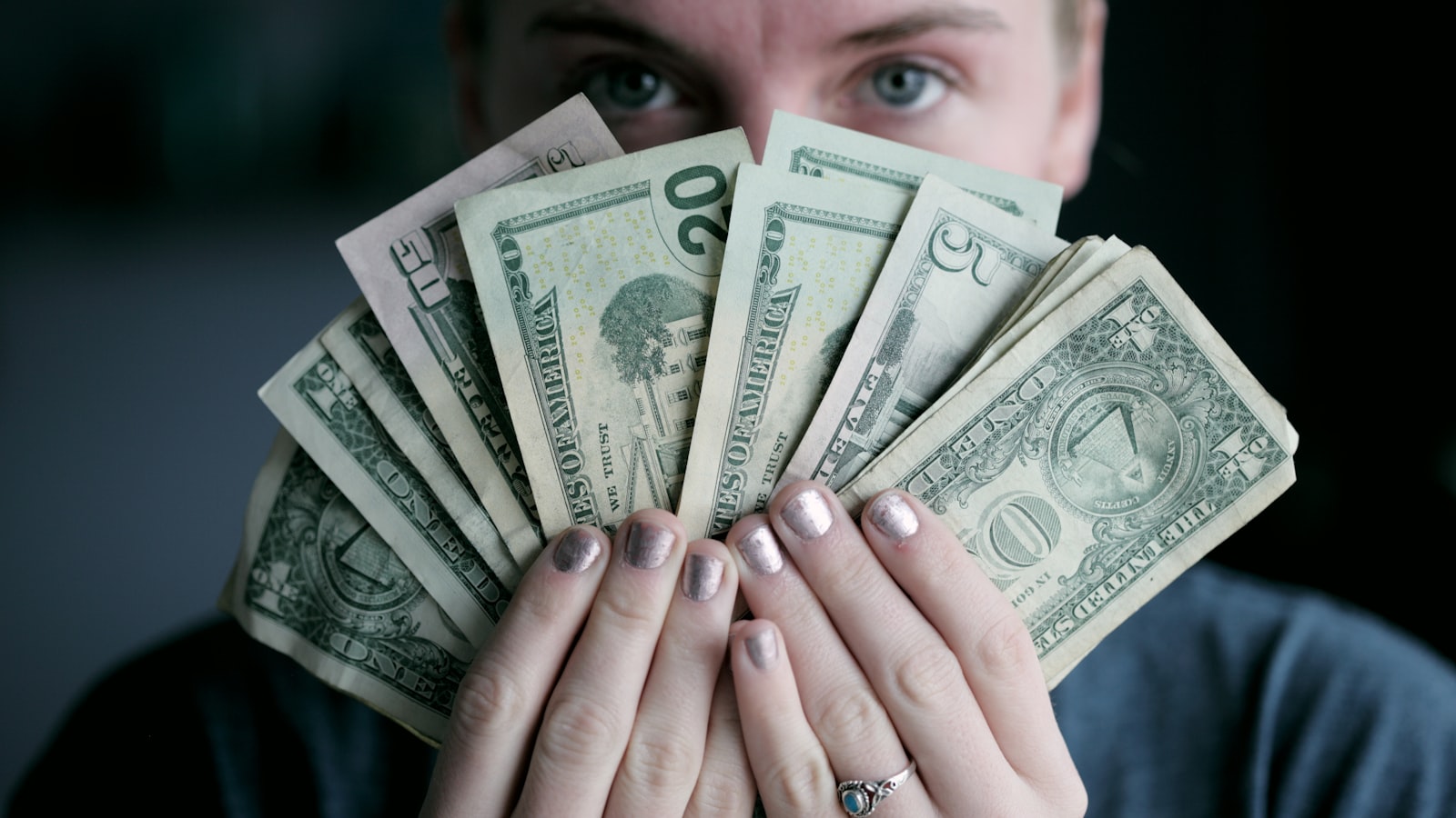 A person holds a fan of US dollar bills in front of their face, revealing only their eyes