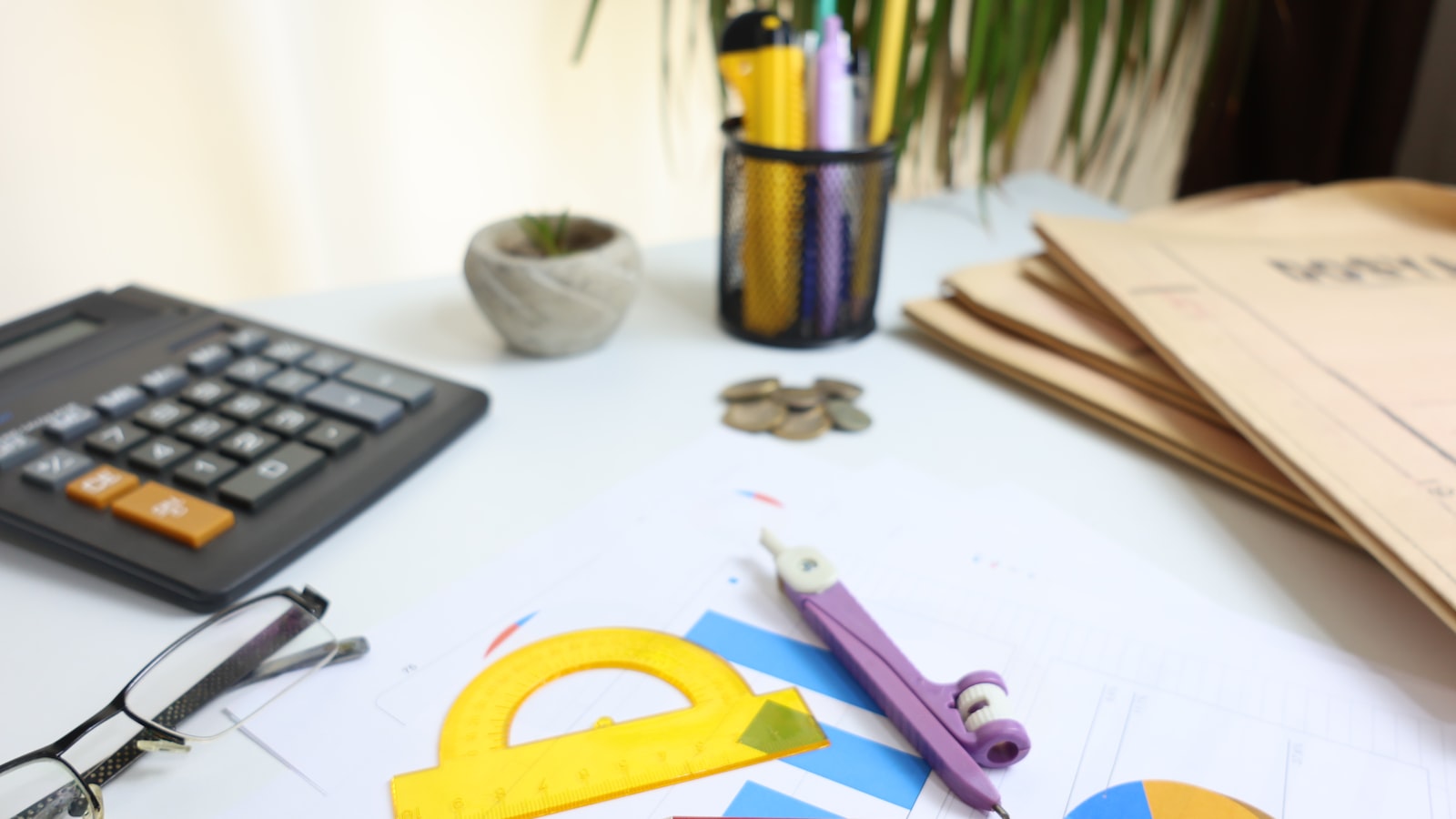 A cluttered desk features a calculator, folders, a pencil cup, coins, glasses, and drafting tools on papers