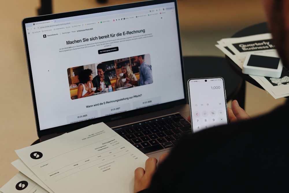 A person holds a phone showing a calculator app, sitting at a desk with a laptop, papers, and a small white device