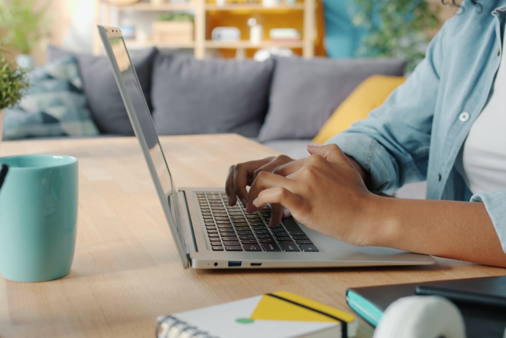 Person typing on a laptop at a wooden table with a blue mug and notebook