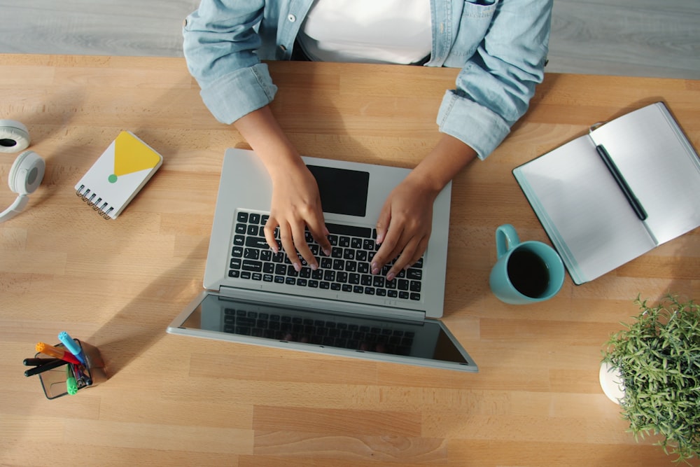 Overhead view of a person typing on a laptop at a wooden desk with notebooks, a mug, pens, and a plant