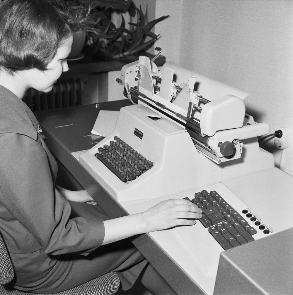 A woman sits at a desk typing on a mechanical accounting machine with a numeric keypad and typewriter keyboard