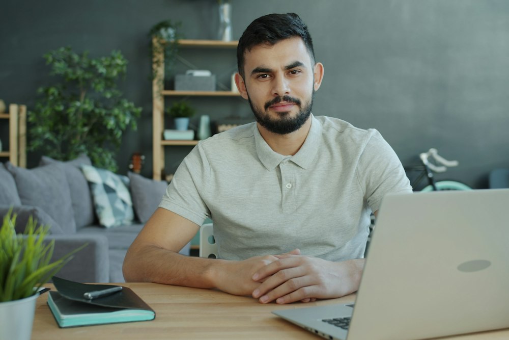 A bearded man in a grey polo shirt sits at a wooden desk with a laptop and notebook, looking forward