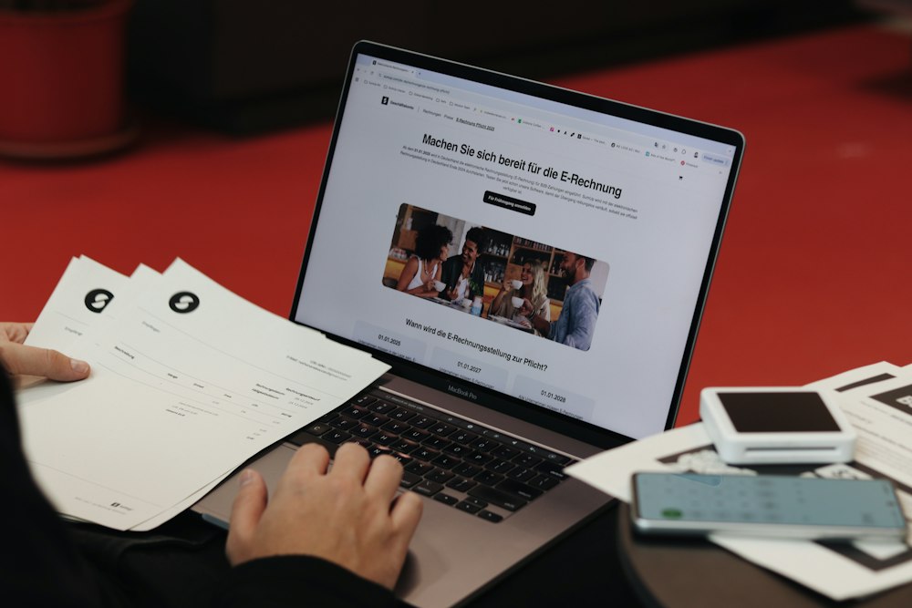 A person holds paper documents while typing on a laptop displaying a website about e-invoices