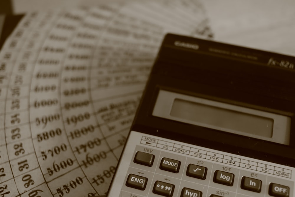 A dark scientific calculator rests on a background of printed numerical charts