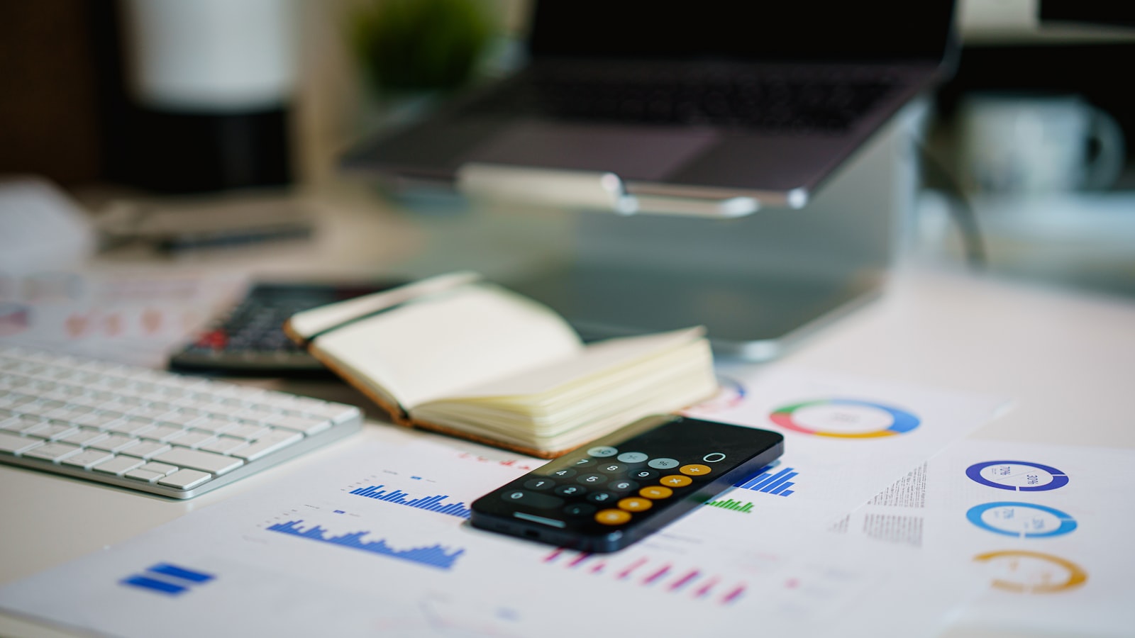 A desk strewn with a keyboard, notebook, smartphone, and financial charts