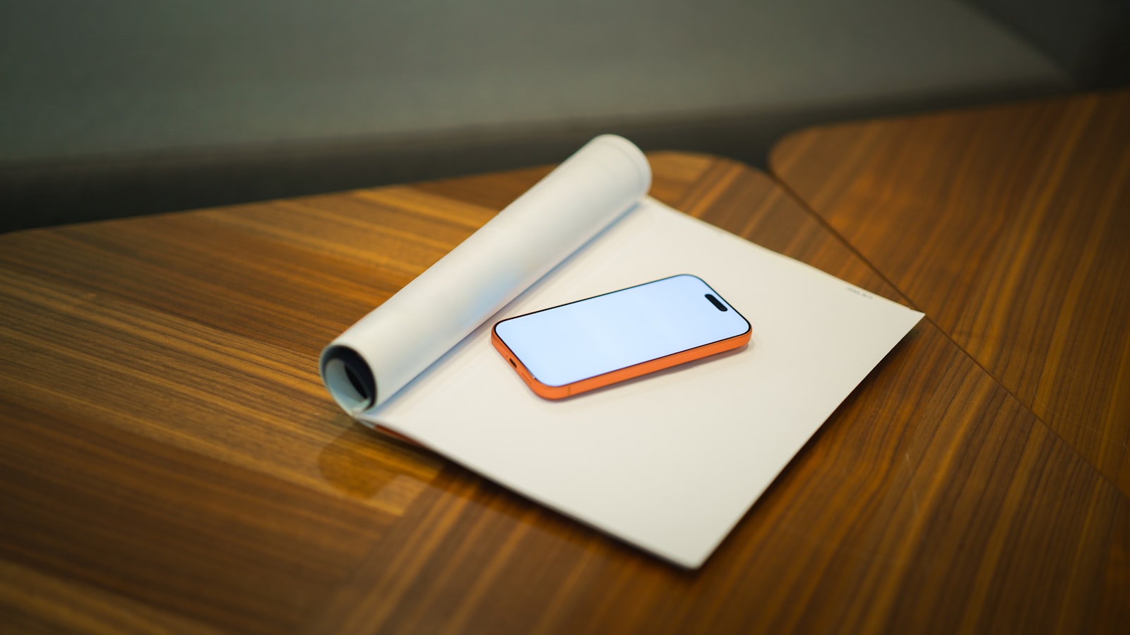 A smartphone with an orange frame and white screen rests on a large white paper pad on a wooden table