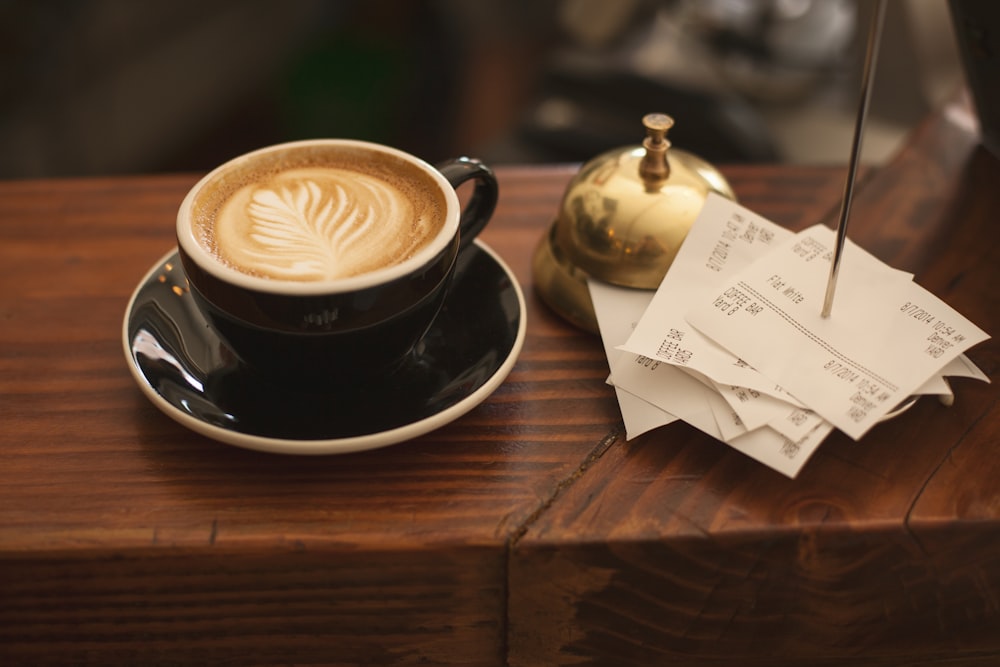 A cup of latte art coffee on a saucer next to a service bell and a stack of receipts on a wooden counter
