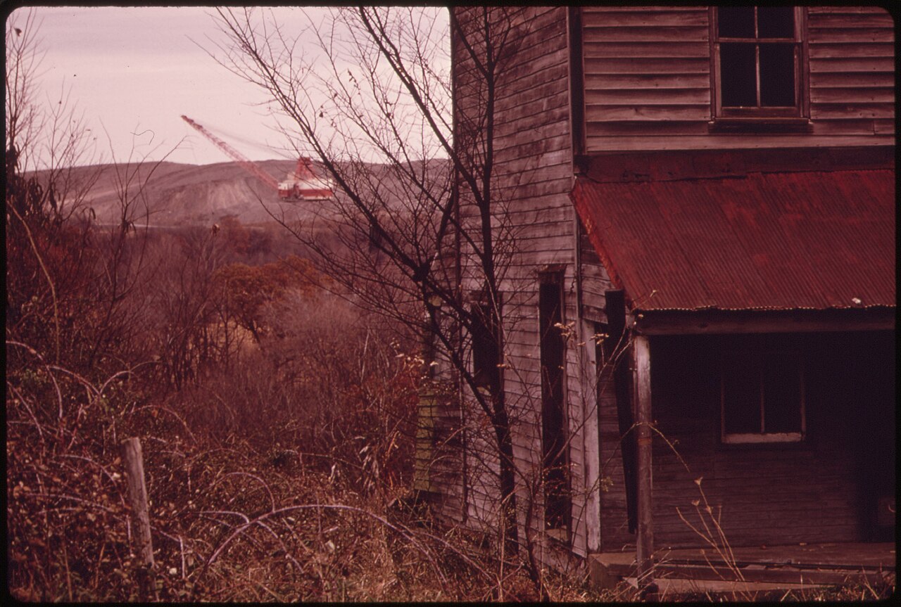 Weathered wooden building with a corrugated red roof and bare trees beside a distant strip mine
