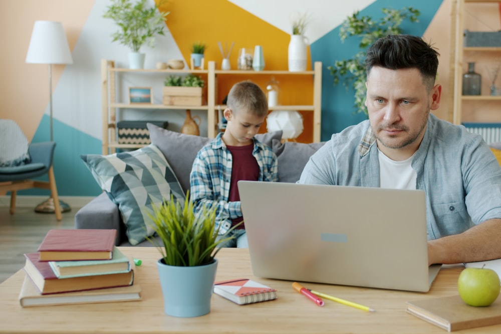 A man works on a laptop at a wooden table while a boy sits on a sofa